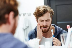 Man brushing his teeth holds side of mouth due to pain