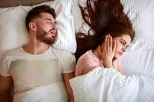 Woman in bed covering ears as her partner snores loudly